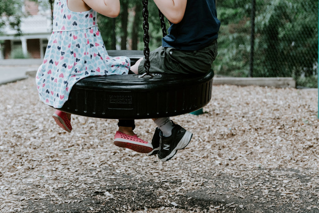 Photograph of two children swinging on a two-seater swing. Only their lower bodies can be seen.