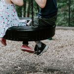 Photograph of two children swinging on a two-seater swing. Only their lower bodies can be seen.