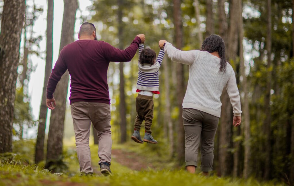 Parents walking in the open woods with a toddler in the middle, and they are holding the toddler up by their hands to lift him off the ground.